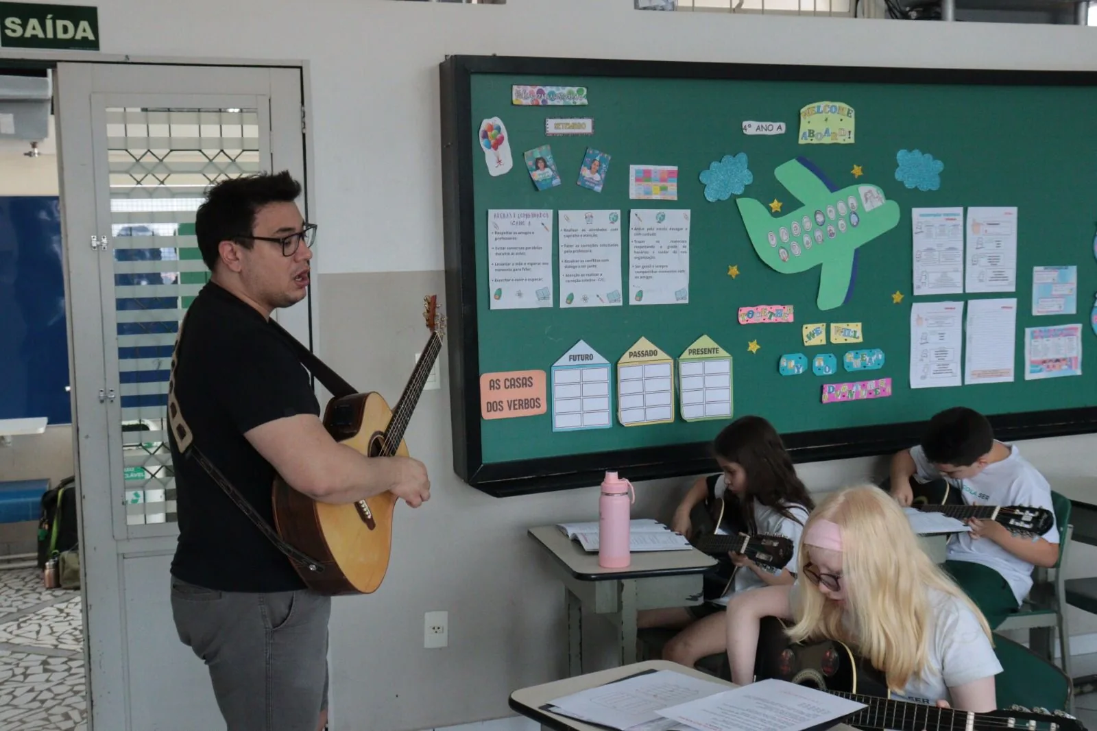 Professor tocando violão em pé durante uma aula em grupo, com os aprendizes sentados acompanhando